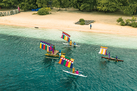 The image depicts an aerial view of Mindanao, Philippines, with five small boats sailing near a sandy beach with lush green trees in the background. Each boat has a brightly colored rectangular sail with various patterns, including stripes and triangles in shades of red, yellow, blue, green, and purple. The clear turquoise water gradually deepens to a darker blue as it stretches away from the shore. One individual is visible walking along the beach, and a set of large letters spelling a word (partially obscured) is positioned on the left side of the sand.